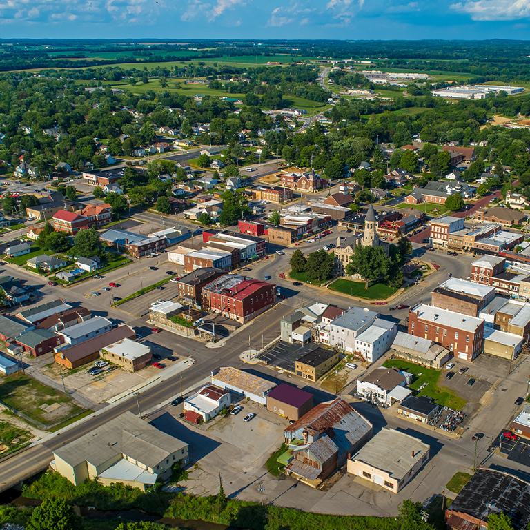 Aerial view of a small Midwestern town with a compact downtown district centered around intersecting streets. Low-rise brick and storefront buildings line the main roads, with a prominent historic church featuring a tall steeple near the center. Surrounding the downtown are residential homes, small businesses, and patches of greenery, transitioning into open farmland and fields in the distance. The scene is brightly lit with blue skies and scattered clouds overhead.