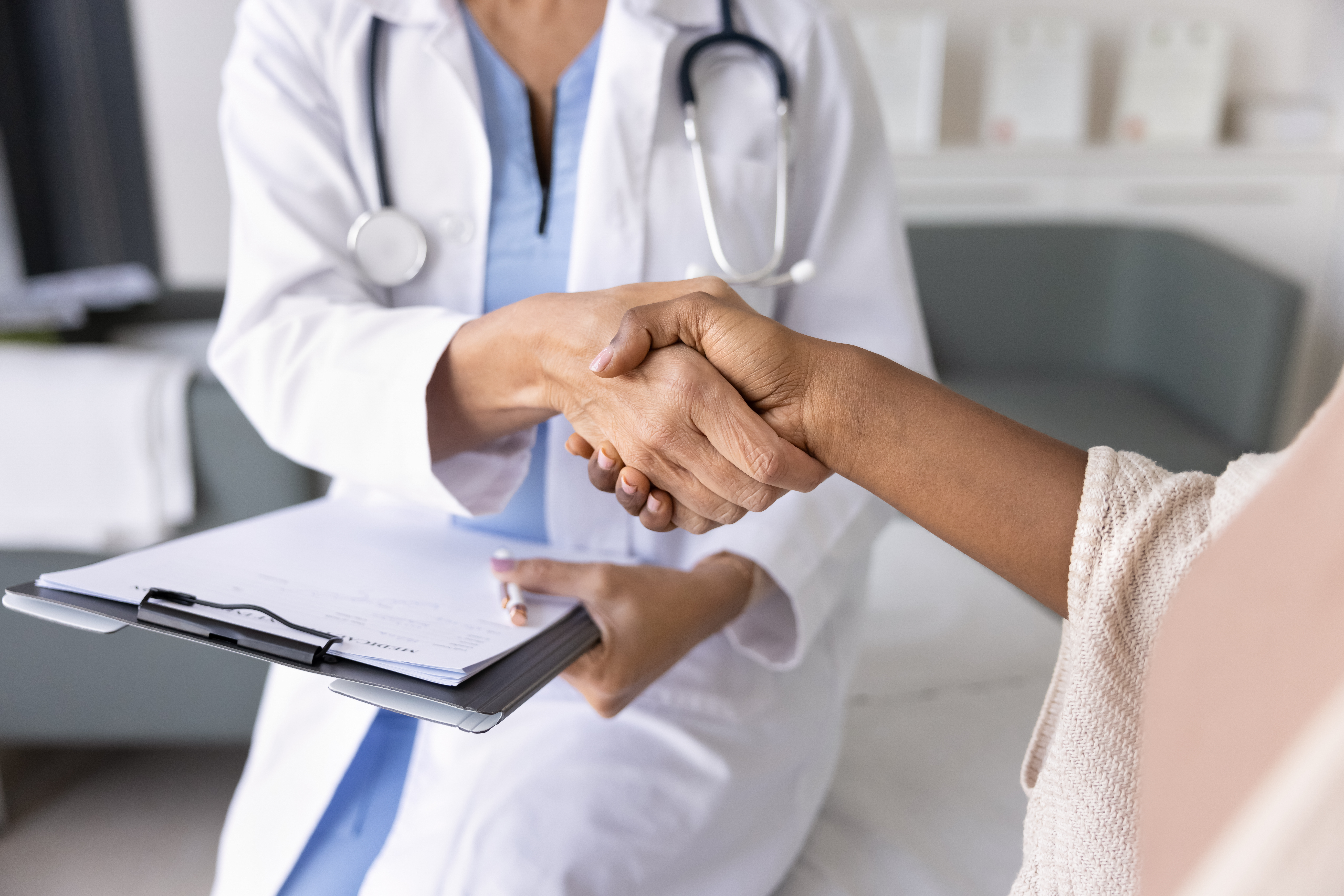 Female doctor holding clipboard and shaking hands with patient; Untitled photo of a doctor in a white coat shakes hands with a patient while holding a clipboard. Image credit: By fizkes, Adobe Stock.; Photo of a doctor in a white coat shakes hands with a patient while holding a clipboard. Image credit: By fizkes, Adobe Stock.